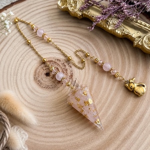 Può includere: Un pendolo con un ciondolo a forma di cono in resina rosa chiaro traslucida con scaglie dorate incorporate. Il ciondolo pende da una catena in metallo dorato decorata con perline rotonde rosa chiaro e piccole sfere dorate, terminando con un ciondolo a forma di gatto dorato.