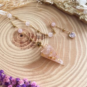 Könnte beinhalten: Ein Rosenquarz-Pendel mit goldfarbener Kette und Akzenten. Das Pendel hat eine dreieckige Form mit Goldeinlagerungen. Die Kette hat kleine rosa Perlen und einen herzförmigen Anhänger mit einem rosa Stein.