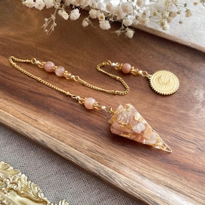 Op de afbeelding: Een goudkleurige pendel met een ketting en een driehoekige kristallen hanger. De hanger is een mix van perzik- en goudtinten, met ingebedde goudvlokken. De ketting heeft perzik-kleurige kralen en een ronde bedel.