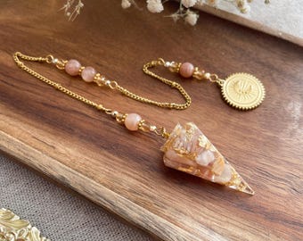 May include: A gold-colored pendulum with a chain and a triangular crystal pendant. The pendant is a mix of peach and gold tones, with gold flakes embedded within. The chain features peach-colored beads and a circular charm.