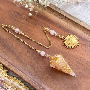 Könnte beinhalten: Ein goldfarbenes Kettenpendel mit einem konischen Anhänger, der mit hellrosa Steinen und Blattgoldflocken gefüllt ist. Die Kette ist mit kleinen, runden, blassrosa Perlen verziert und endet in einem herzförmigen, goldfarbenen Anhänger mit Augenmotiv.