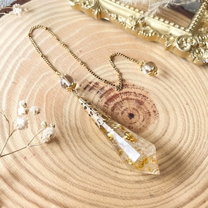 Könnte beinhalten: Ein goldfarbenes Pendel mit einem klaren, facettierten Kristallanhänger, der mit Goldflocken gefüllt ist. Das Pendel hat eine zarte Kette und eine kleine Kristallperle an der Spitze. Der Hintergrund ist eine Holzoberfläche mit einem dekorativen Goldrahmen.