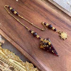 Könnte beinhalten: Ein Pendel mit einer konischen Harzspitze, gefüllt mit dunkelroten und goldenen Flocken, aufgehängt an einer goldfarbenen Kette. Die Kette ist mit dunkelroten Perlen und einem goldfarbenen Sonnenanhänger am Ende verziert, ein dekoratives Stück.