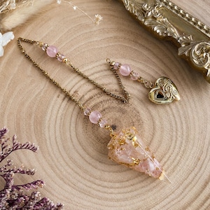 Peut inclure: Un pendule composé d'un cristal de quartz rose en forme de cône incrusté de flocons dorés, suspendu à une chaîne dorée. La chaîne est ornée de petites perles roses et d'un médaillon doré en forme de cœur avec un motif floral en relief.