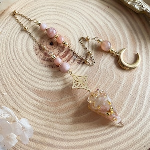 May include: A gold-colored pendulum with a clear, cone-shaped end filled with pink and gold flakes. The pendulum is attached to a gold chain with pink gemstone beads and a gold crescent moon charm. The design is on a wooden surface.