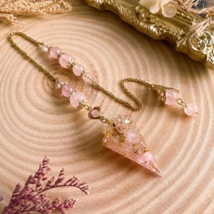May include: A pendulum featuring a cone-shaped pendant made of pink resin with gold flakes, attached to a gold-tone chain. The chain includes several round pink beads, a small heart-shaped pink charm, and a decorative gold-tone metal cap.