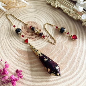 Op de afbeelding: Een goudkleurige pendel met een donker bordeauxrode, facetgeslepen kristal en accenten van bladgoud. De ketting heeft zwarte en rode kralen en een rood hartvormig bedeltje. De pendel wordt gepresenteerd op een houten oppervlak.