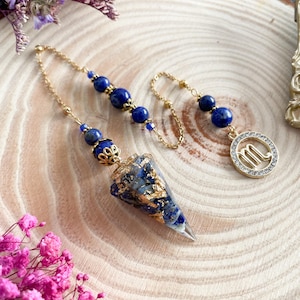 Peut inclure: Un pendule à chaîne dorée avec des perles bleues et une extrémité pointue transparente. Le pendule comporte un charm doré avec le symbole du zodiaque Scorpion. Le pendule est décoré de paillettes dorées.