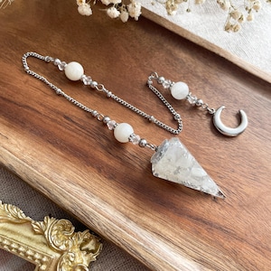 Peut inclure: Un pendule argenté avec une pointe en cristal triangulaire transparente et une chaîne ornée de perles blanches. Une chaîne séparée avec une perle blanche et un croissant de lune argenté est également présente. Les articles sont présentés sur un plateau en bois.