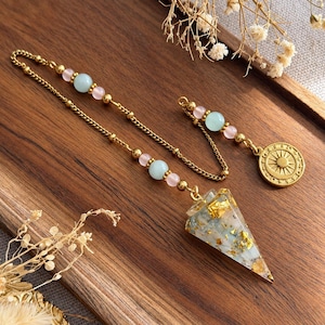 May include: A gold-toned metal chain pendulum featuring a conical resin point filled with gold flakes. The chain is accented with alternating light blue and pale pink spherical beads, ending in a circular gold-toned charm with a sunburst design.
