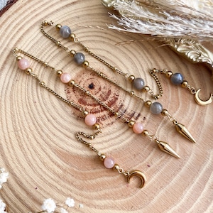 Peut inclure: Boucles d'oreilles en chaîne dorée avec des perles roses et grises, et des breloques en forme de croissant de lune et de pointe dorées. Présentées sur une surface en bois, elles mettent en valeur leur design délicat.