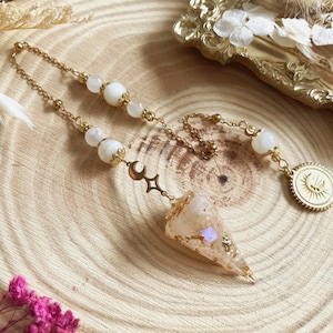 Può includere: Un pendolo color oro con catena e perline bianche. Il pendolo ha una punta di cristallo con scaglie d'oro e un ciondolo a forma di luna e stella. È attaccata anche una moneta d'oro con un disegno del sole.