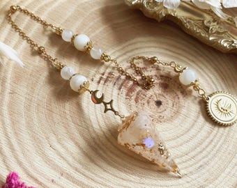 May include: A gold-colored pendulum with a chain and white beads. The pendulum has a crystal point with gold flakes and a moon and star charm. A gold coin with a sun design is also attached.