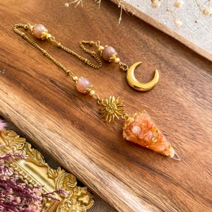 Può includere: Un pendolo dorato con un ciondolo a forma di sole, una punta di cristallo trasparente e arancione e una catena con perline rosa. È presente anche un ciondolo a forma di mezzaluna dorata. Gli oggetti sono esposti su una superficie di legno.