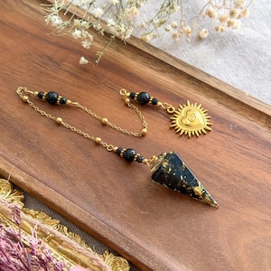 Könnte beinhalten: Ein Pendel mit einem schwarzen, gold gesprenkelten konischen Stein, der an einer goldfarbenen Kette hängt. Die Kette ist mit kleinen schwarzen Perlen verziert und endet in einem goldfarbenen Anhänger in Herzform mit einem Auge und Strahlen.