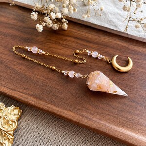 May include: A pendulum featuring a pointed, translucent pink resin crystal with gold leaf flakes, attached to a gold-tone chain. The chain includes small, round, pale pink beads and terminates in a polished gold-tone crescent moon charm.