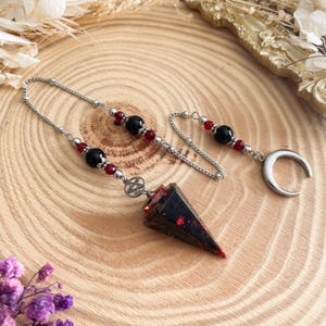 Peut inclure: Un pendule avec une pointe en cristal noir et rouge, une chaîne argentée et des perles décoratives. La chaîne est ornée de perles noires et rouges, et d'un croissant de lune. Un charme de pentagramme est attaché à la pointe du cristal.