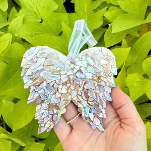 May include: An angel wing decoration made from iridescent mosaic tiles. The wings are attached to a sheer ribbon for hanging. The ornament is held against a backdrop of green leaves.