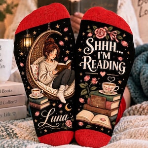 Op de afbeelding: Een paar zwarte sokken met rode boorden, voorzien van een afbeelding van een lezend persoon in een hangstoel en de tekst SHHH... I'M READING boven een stapel boeken en rozen, met de naam Luna in witte letters op de sok gedrukt.