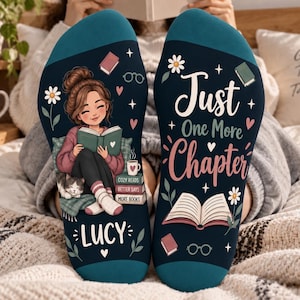 May include: A pair of dark teal socks featuring a graphic of a person reading a book with a cat, the name Lucy, and the text Just One More Chapter, accompanied by illustrations of books, flowers, and glasses on a soft fabric background.