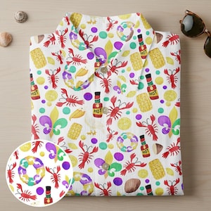 May include: White button-down shirt with a colorful pattern of red crawfish, hot sauce bottles, corn, and purple fleur-de-lis. The shirt features a small, circular pattern of the same design. Sunglasses and seashells are in the background.