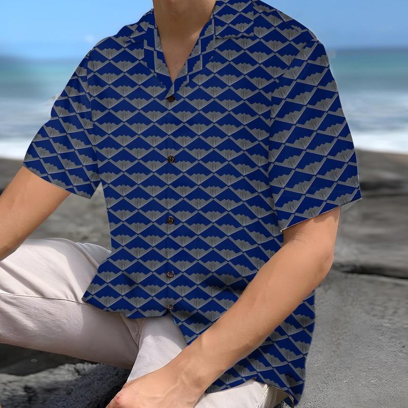 Puede incluir: Una camisa de manga corta con un patr&oacute;n geom&eacute;trico azul y gris. La camisa tiene un cuello cl&aacute;sico y botones de madera. El patr&oacute;n es un dise&ntilde;o repetitivo de formas grises sobre un fondo azul. La camisa est&aacute; hecha de tela ligera.