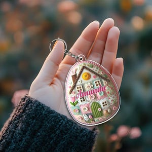May include: A personalized keychain featuring a house design with the name "Amanda" in pink. The keychain has a silver ring and chain. The house design includes a chimney, windows, and a green door, with decorative buttons.