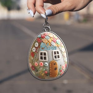 May include: An oval-shaped keychain with a miniature house design. The house has a grey facade, two windows, and a brown door. It is decorated with colourful flowers, buttons, and the name "Olivia" in cursive. The keychain has a silver-coloured metal border.