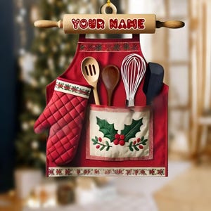 May include: A red apron-shaped Christmas ornament with a rolling pin at the top that reads "YOUR NAME". The ornament features a red oven mitt, wooden spoon, whisk, and spatula. A pocket on the apron has a holly design.