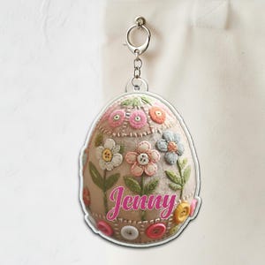 May include: An egg-shaped keychain with a floral design. The keychain features a beige background with colorful felt flowers and green leaves. The name "Jenny" is written in pink script. The keychain is attached to a silver metal clasp.