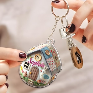 Peut inclure: Un porte-clés en forme de maison avec un design patchwork, avec des carrés de tissu floral et uni, des boutons en bois et un petit lapin. Le nom "Sophia" est écrit en rose. Comprend une étiquette en bois avec une icône de maison.