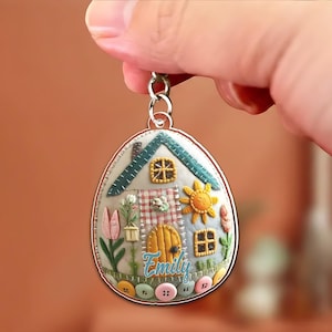 May include: A handmade keychain featuring a whimsical house design. The egg-shaped pendant showcases a miniature house with a green roof, a yellow door, and a sun. The name "Emily" is written in cursive. Colorful buttons and floral accents complete the design.