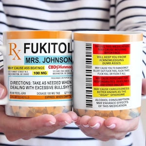 May include: Two orange and white mugs with a humorous prescription label design. The text on the mugs reads "RX FUKITOL" and "MAY CAUSE ASS BEATINGS". The mugs are held by a person's hands.