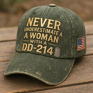 May include: Distressed olive green baseball cap with the embroidered text "NEVER UNDERESTIMATE A WOMAN WITH A DD-214" in gold. Features a small American flag patch on the side and a dog tag graphic. The cap has a curved brim.