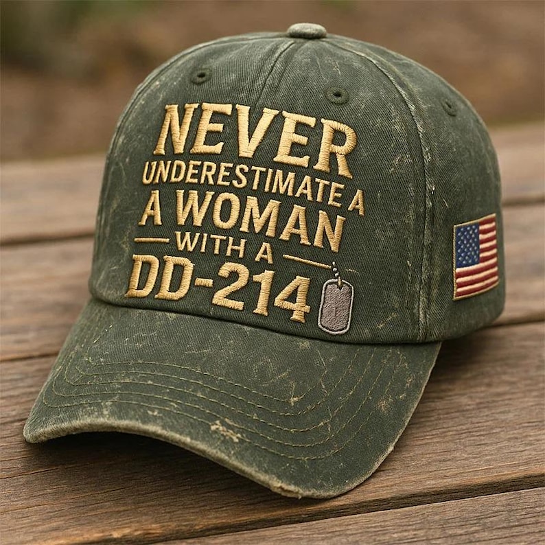 May include: Distressed olive green baseball cap with the embroidered text "NEVER UNDERESTIMATE A WOMAN WITH A DD-214" in gold. Features a small American flag patch on the side and a dog tag graphic. The cap has a curved brim.