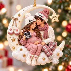 May include: A decorative Christmas ornament featuring a couple sitting on a crescent moon. The person on the left wears a white beanie labeled Ava and holds an ultrasound image. The person on the right wears a pink beanie labeled Lucas. A star reads 2026.
