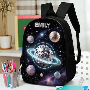 May include: Black backpack with a space-themed design featuring planets, stars, and a glowing ring around a large planet. The name "EMILY" is printed at the top. The backpack has black straps and a zipper.