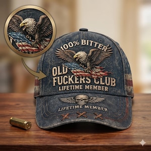 May include: A distressed blue baseball cap with an eagle graphic holding an American flag. The cap features the text "100% Bitter" and "Old Fuckers Club Lifetime Member". A gold-colored bullet casing is on the wooden surface.