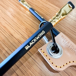 May include: Two black pencils with gold lettering that reads "Blackwing" and "Blackwing" on the side. The pencils are attached to a brown leather keychain with a metal ring.