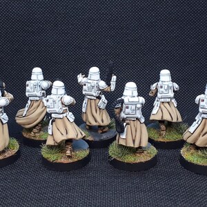 Op de afbeelding: Een groep van zeven wit en bruin geverfde miniatuurfiguren die witte helmen dragen en wapens dragen. De figuren staan op ronde bases met groen gras.