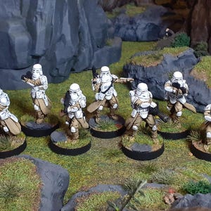 Op de afbeelding: Een groep van zeven wit en bruin geverfde miniatuurfiguren van Star Wars Stormtroopers, die elk op een ronde zwarte basis staan met groen gras. De figuren houden wapens vast en staan in verschillende actiehoudings.
