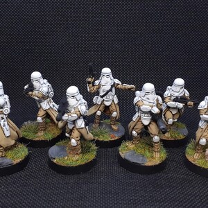 Op de afbeelding: Een groep van zeven wit en bruin geschilderde miniatuurfiguren van Star Wars Stormtroopers. De figuren staan in verschillende actiehoudings, houden wapens vast en staan op ronde bases.