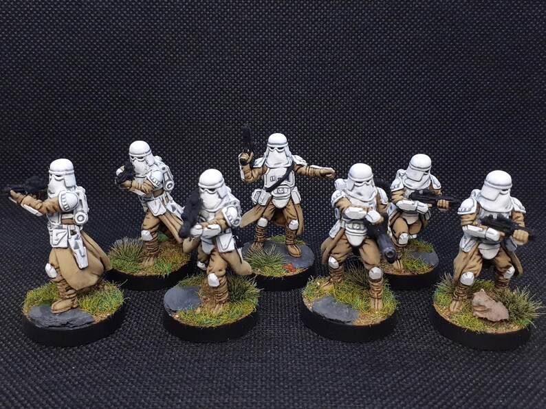 Op de afbeelding: Een groep van zeven wit en bruin geschilderde miniatuurfiguren van Star Wars Stormtroopers. De figuren staan in verschillende actiehoudings, houden wapens vast en staan op ronde bases.