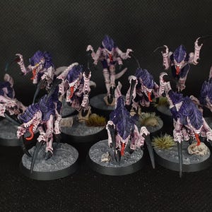 Puede incluir: Un conjunto de figuras en miniatura que representan criaturas insectoides con armadura púrpura y rosa, garras afiladas y lenguas rojas. Las figuras están montadas sobre bases grises, lo que sugiere un juego de mesa o un hobby.