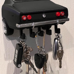 Può includere: Un portachiavi da parete a forma di parte posteriore di un'auto vintage nero opaco. Presenta un paraurti color argento, quattro luci posteriori circolari rosse e cinque ganci neri sottostanti per appendere portachiavi, telecomandi e chiavi.