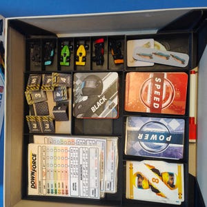 May include: A game box containing a black and white game board, cards with the words "Speed", "Power", and "Downforce", and small black, green, and red racing cars.