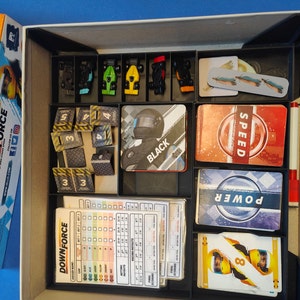 May include: A game box organiser with compartments for game pieces, cards, and instructions. The organiser is black with a grey interior. The game pieces include small cars, numbered tiles, and cards with the words "Speed", "Power", and "Downforce".