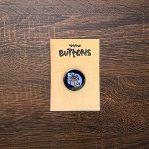 May include: A round button with a cartoon image of a character holding a sign that says "F*ck ICE". The button is on a tan card that says "tiny buttons" in black letters. The background is a dark brown wood.