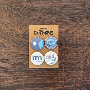 May include: A set of four circular pin-back buttons attached to a brown cardboard display card labeled tinyino BUTTONS. The buttons feature designs including a blue Minnesota flag, the state seal, the letters mn, and a Minnesota State Fair graphic.