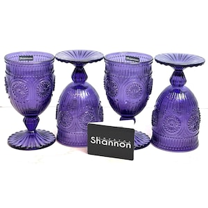 May include: Four purple glass goblets featuring a pressed floral pattern and vertical ribbing. Two glasses stand upright, while two are inverted. A black rectangular card with the white text Shannon Crystal is positioned in the center foreground.